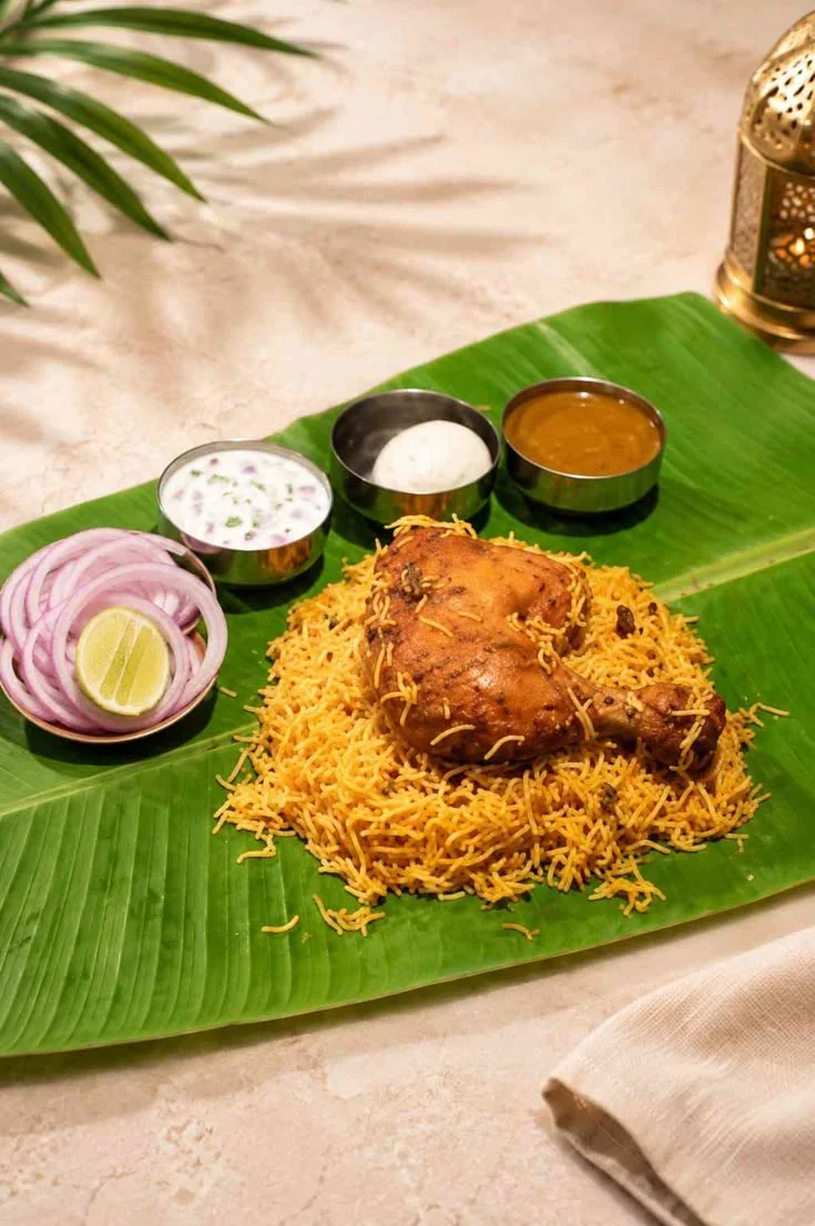 A vibrant Hyderabadi chicken biryani served on a banana leaf with raita, curry, and onions